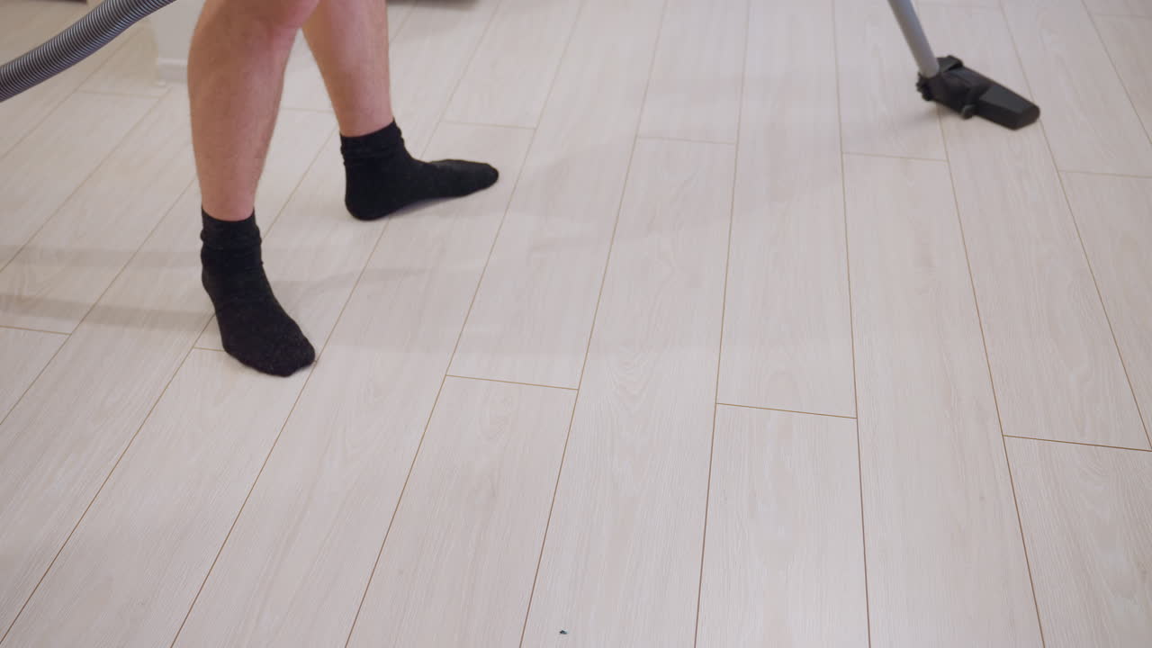 Close up of man vacuuming light wooden floor with modern cleaner, black socks visible while standing on clean surface, concept of household chores, hygiene maintenance, and daily home cleaning