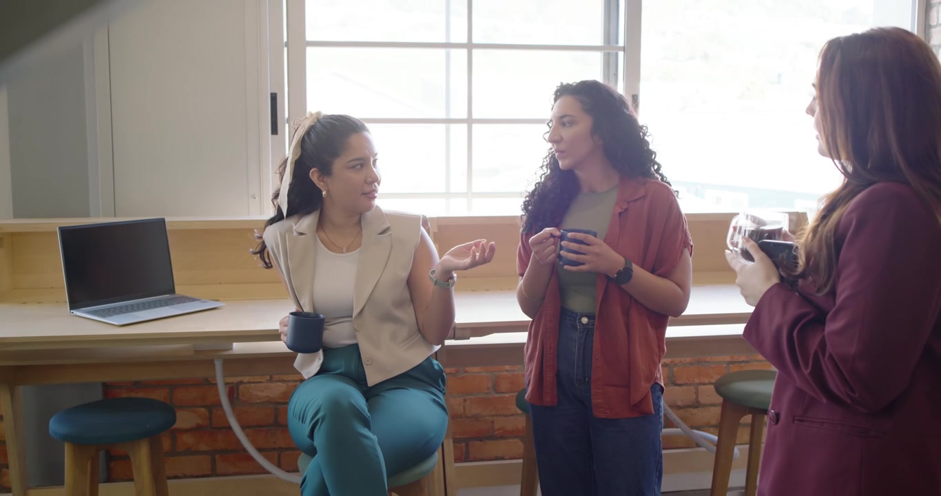 Three women coworkers talking at office bar on break while right woman gesturing, mugs and laptop
