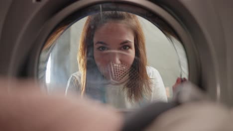 View-from-inside-the-washing-machine-portrait-of-a-happy-brunette-girl-in-a-white-T-shirt-who-loads-things-into-the-washing-machine-and-looks-into-it-from-the-outside-in-a-modern-apartment