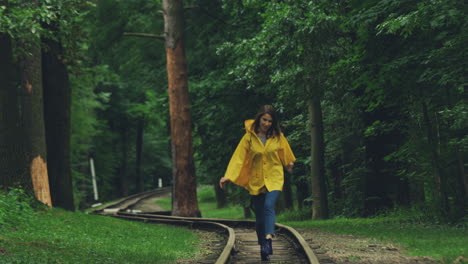 una joven alegre con un impermeable amarillo corriendo alegremente en el antiguo ferrocarril durante la tormenta