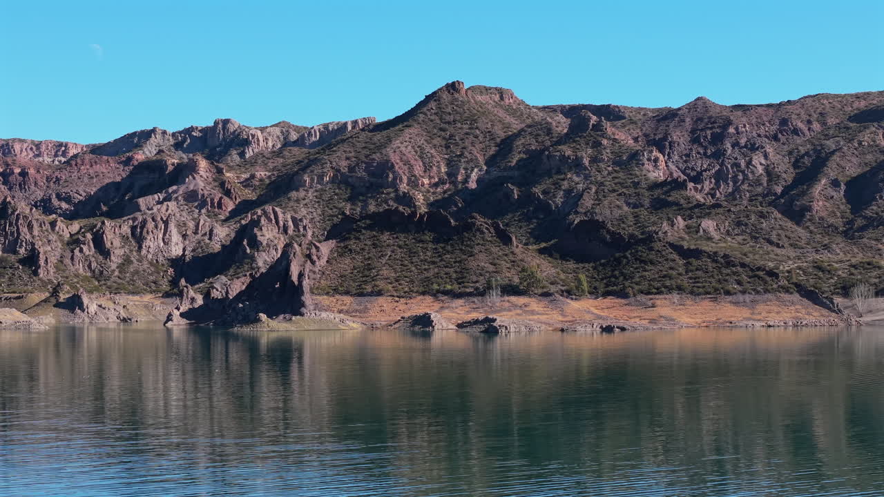 Drone shot captures rugged reddish cliffs reflected on the still waters of the Atuel River reservoir in Cañón del Atuel, San Rafael, Mendoza, Argentina, under a clear sky, real time