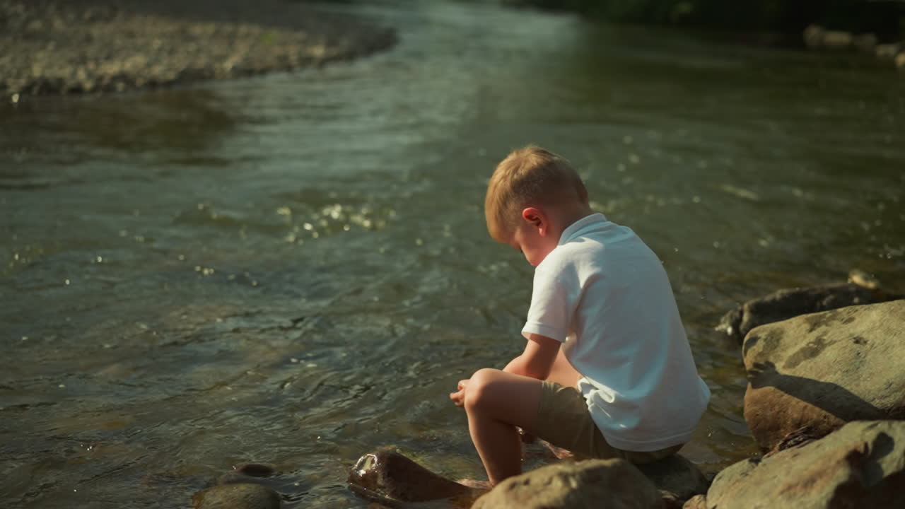 Blond boy sits on rock and throws stones into water. Little child in white t-shirt plays by shallow river. Concept of active lifestyle on warm summer day