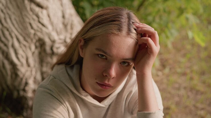 Young woman seated on ground rests her head on her left hand, gazing up thoughtfully with a contemplative expression, with background featuring tree bark, foliage, and greenery