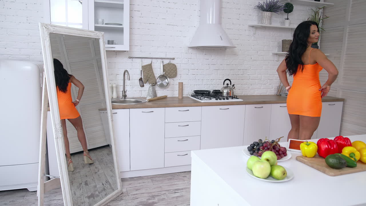 Confident brunette woman standing in front of the mirror and looking at her reflection while preparing breakfast at the kitchen. Stock photo