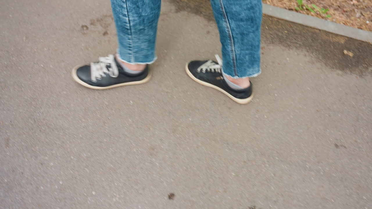 Black Sneakers On Pavement Walking Denim Legs CloseUp Capturing Steady Stride Along Urban Sidewalk, Visible Laces And Rubber Soles, Relaxed Tempo, Ankle Motion, Neutral Asphalt Surface, Everyday
