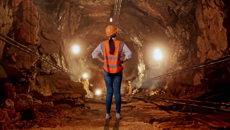 full body back view of a female engineer wearing safety helmet looking around while standing with arms akimbo in underground mine tunnel