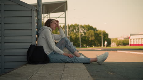 girl in jeans seated on ground, resting hand on head with thoughtful expression, black bag nearby, background includes blurred figure walking and distant building