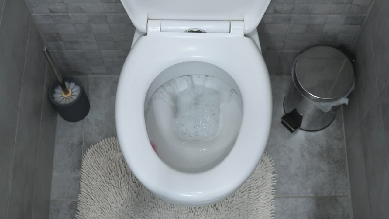 A top-down view of a toilet being flushed in a modern bathroom. The scene includes a toilet brush, a small trash bin, and a textured bath mat, creating a clean and organized space.