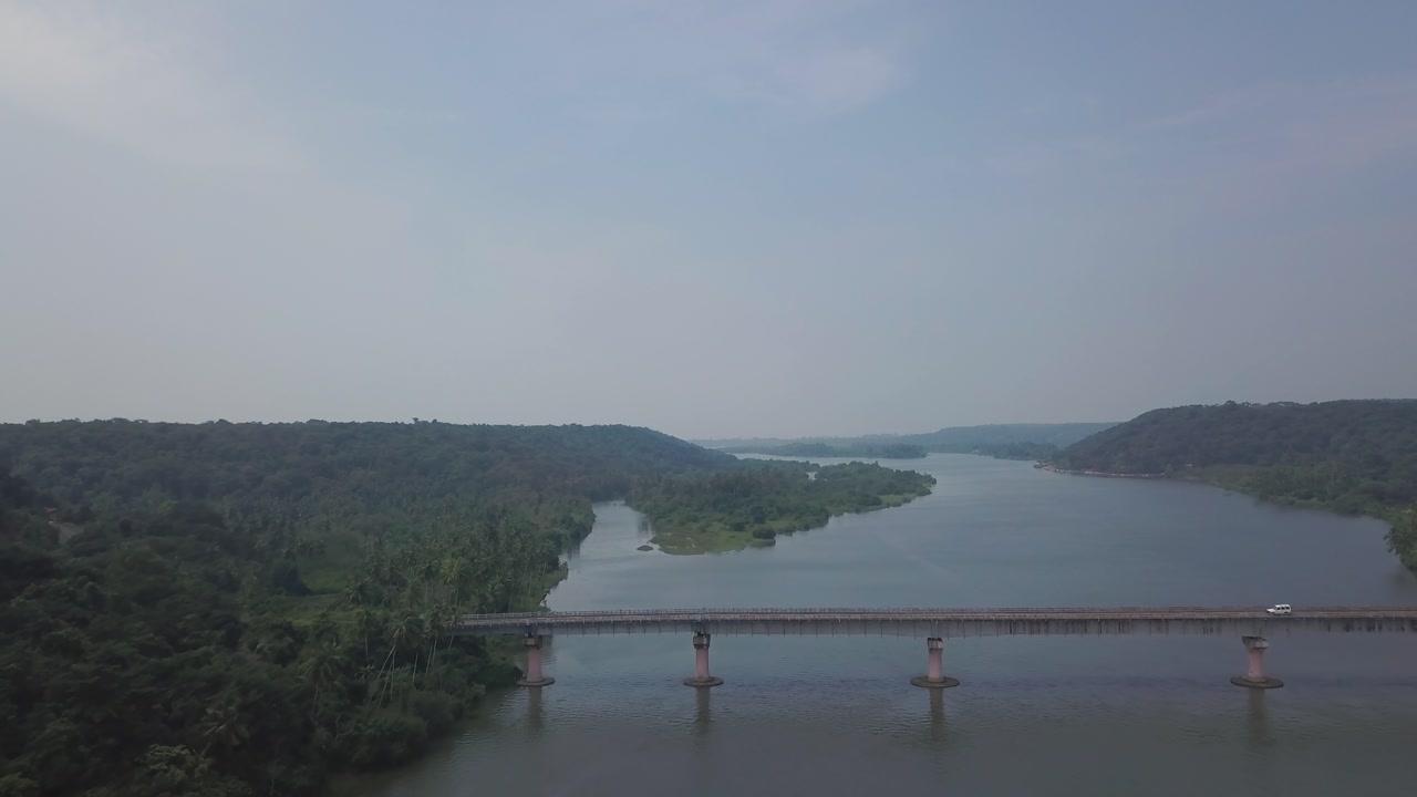 Concrete bridge spanning calm backwaters of Karli River near Devbag and Malvan in Maharashtra, India. Green forested hills line both riverbanks under overcast sky.