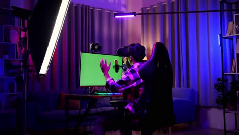 back view of asian boy streamer playing game, touching, and swiping experience virtual reality in futuristic goggles. green screen personal computer on desk illuminated by rgb led strip light