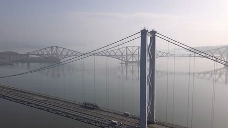 Aerial-footage-of-the-old-Forth-Road-Bridge-with-the-Forth-Railway-Bridge-in-the-background-on-a-sunny-day-at-South-Queensferry-in-West-Lothian,-Scotland