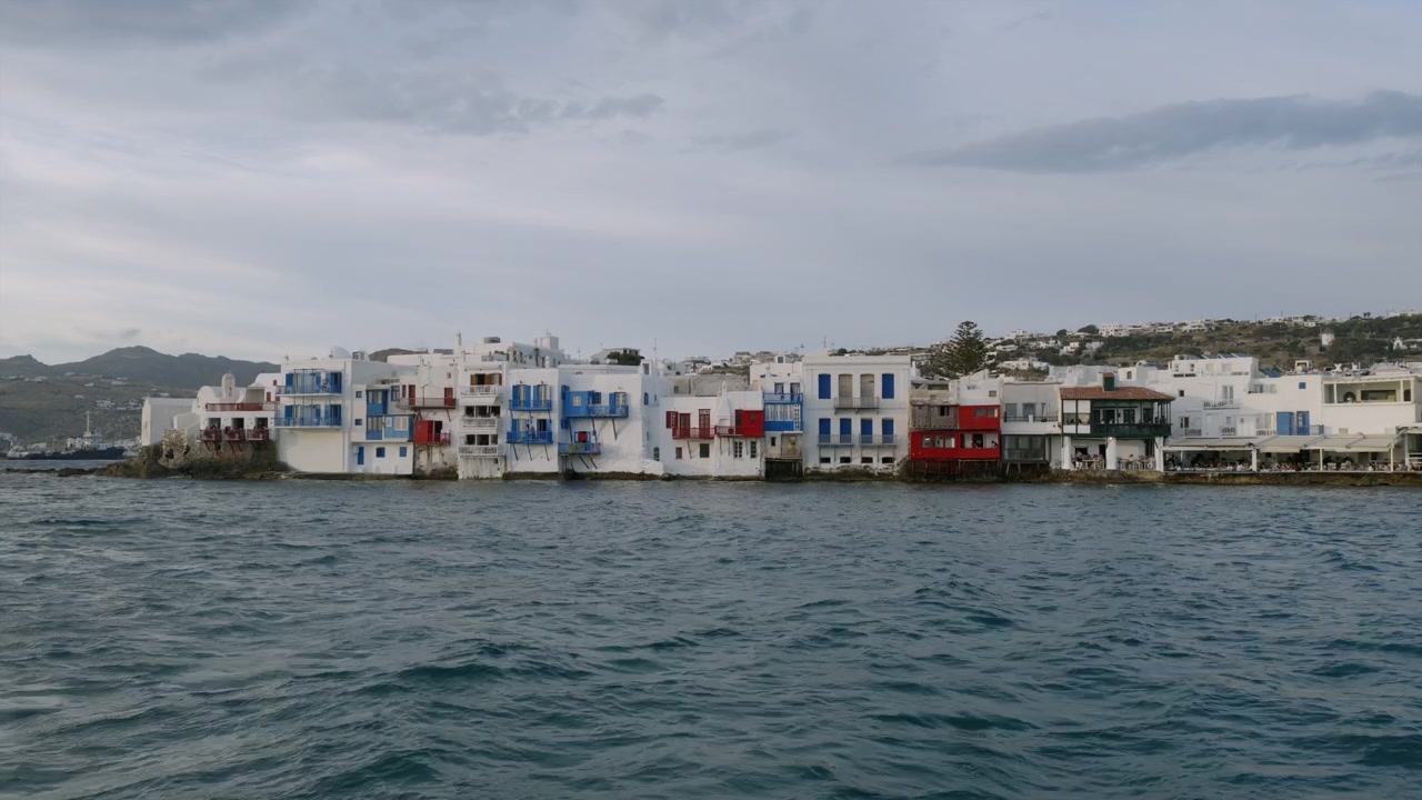 Lungomare di Piccola Venezia a Mykonos sotto un cielo nuvoloso e tenue, con case bianche e balconi colorati che sorgono dal mare, visto dalla riva.