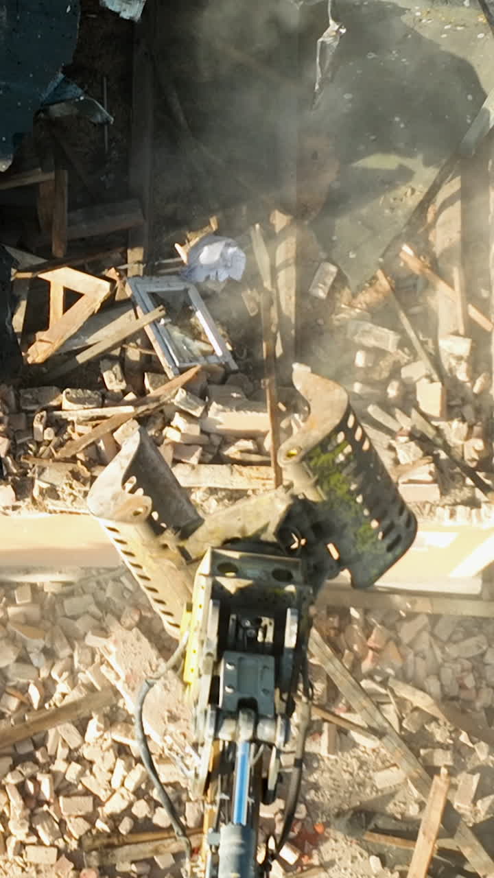 A highangle aerial shot shows a hydraulic excavator claw tearing through a brick wall and wooden debris during a building demolition. Useful for construction, urban renewal, and waste management conte
