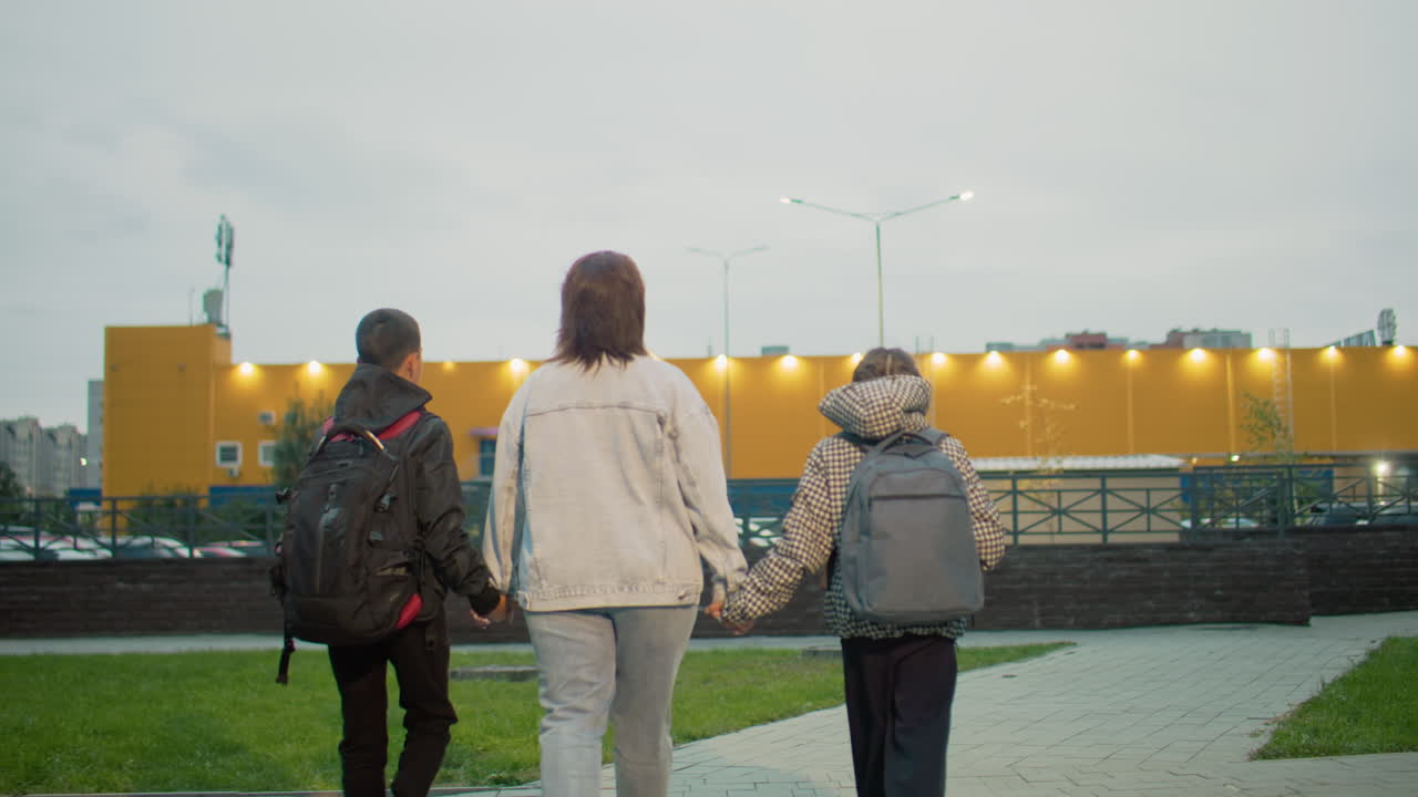 mum walking kids hand in hand on urban park pathway at dusk wearing backpacks strolling near playground against city backdrop capturing family support evening routine indulgent care