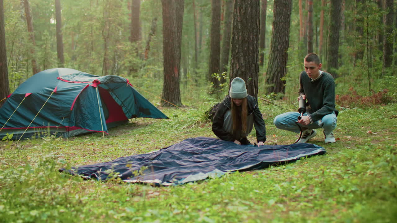 Young couple spending time together in forest campsite as man pumps air into tent bag while woman looks at him warmly, surrounded by nature and glowing morning light near pitched camping tent