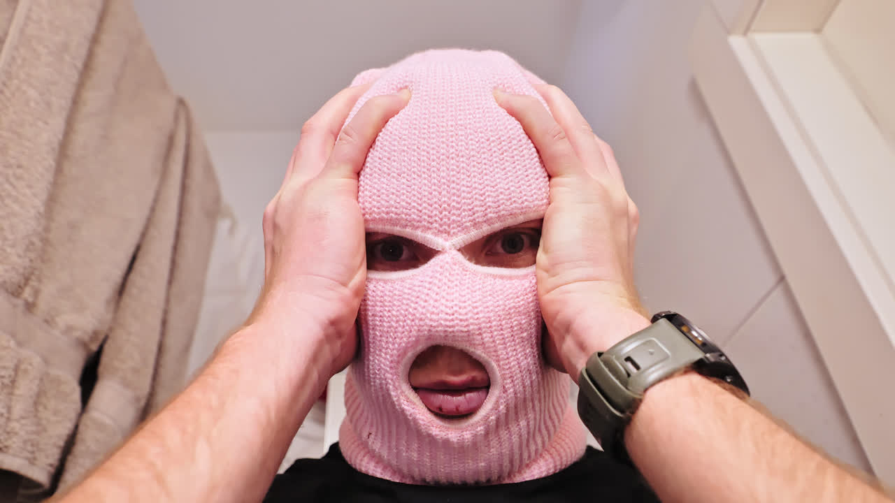 Masked man in pink balaclava nervously holds head in panic in bright bathroom