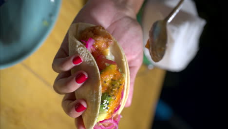 vertical slow motion of a female woman holding a mexican traditional taco with avocado and red onion, pouring red hot salsa on it