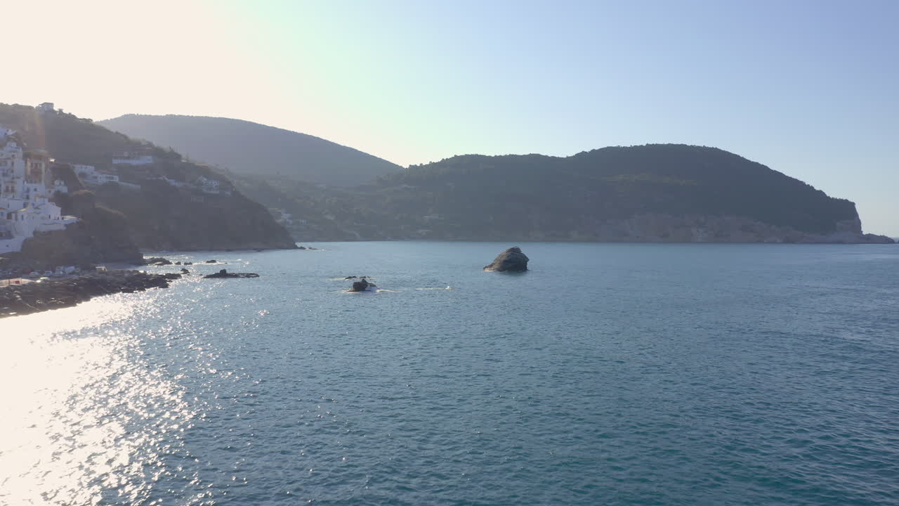 Aerial: Passing over a big rock in the middle of the blue clear and tranquil sea near Skopelos island harbor