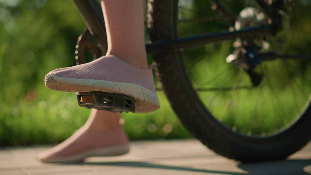 primer plano de la pierna en zapatillas de deporte tocando el pedal de la bicicleta mientras el otro pie descansa en el suelo, la bicicleta se desplaza ligeramente hacia adelante mientras el ciclista se equilibra, contra la vegetación del parque en un fondo borroso