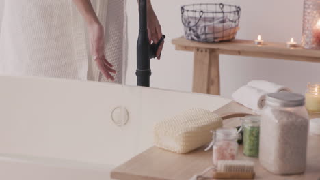 la cámara enfoca las manos de una mujer que lleva un albornoz abriendo un grifo para llenar la bañera con agua