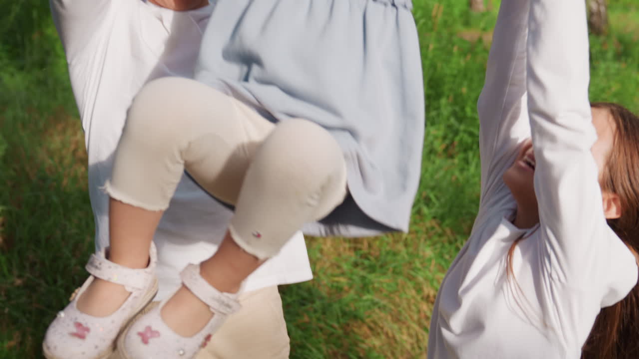 Aerial view of brother and sisters walking on forest path holding hands, gently lifting youngest sibling between them, surrounded by bright sunlight, creating joyful outdoor family bonding moment