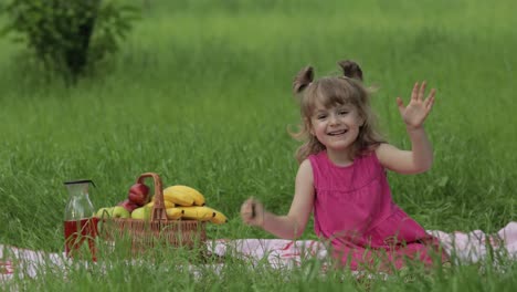 Weekend-at-picnic.-Lovely-caucasian-child-girl-on-green-grass-meadow-sit-on-blanket-waving-her-hands