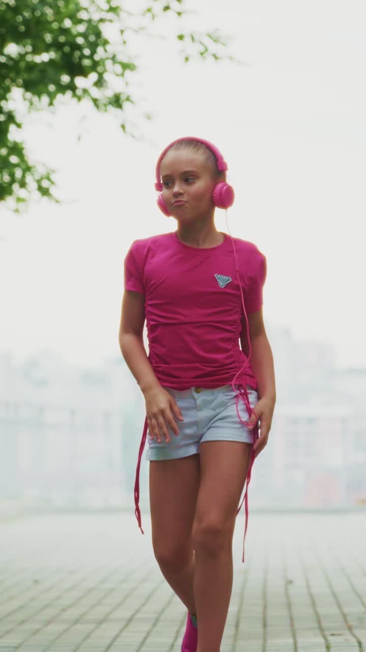 Ein junges Mädchen in einem pinken Shirt tanzt draußen mit Kopfhörern und fängt einen freudigen Moment ein, während sie sich auf einem sonnigen, von Grün umgebenen Weg zur Musik bewegt.