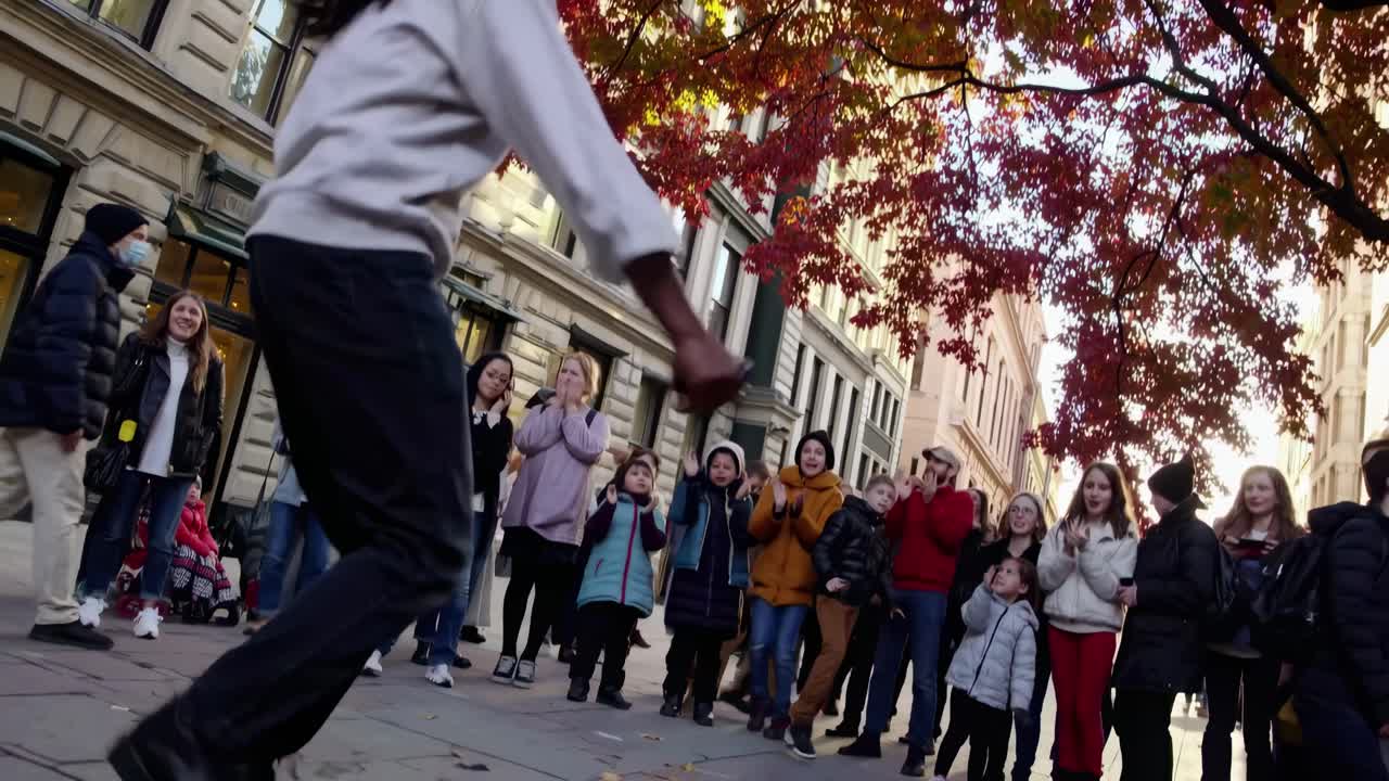 Street performer dances energetically, captured from a low angle. A crowd watches, framed by autumn