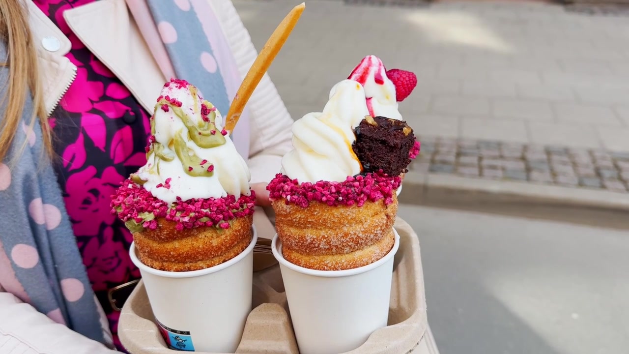 Twee kleurrijke schoorsteencake desserts in afhaalbekers, getopt met softijs, frambozenkruimels, brownie, aardbei, pistachesaus en wafelstokjes