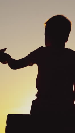 boy is holding paper ship in his hand and playing with it on the background of sunset by the river in the summer. Vertical video