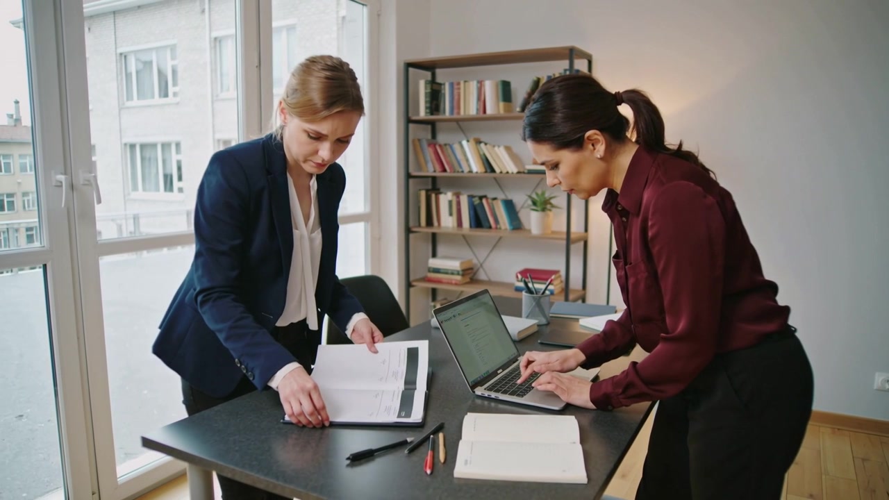 Donne che aprono un raccoglitore, sistemano documenti in ufficio, digitano su un laptop, preparano un rapporto, in abbigliamento business casual
