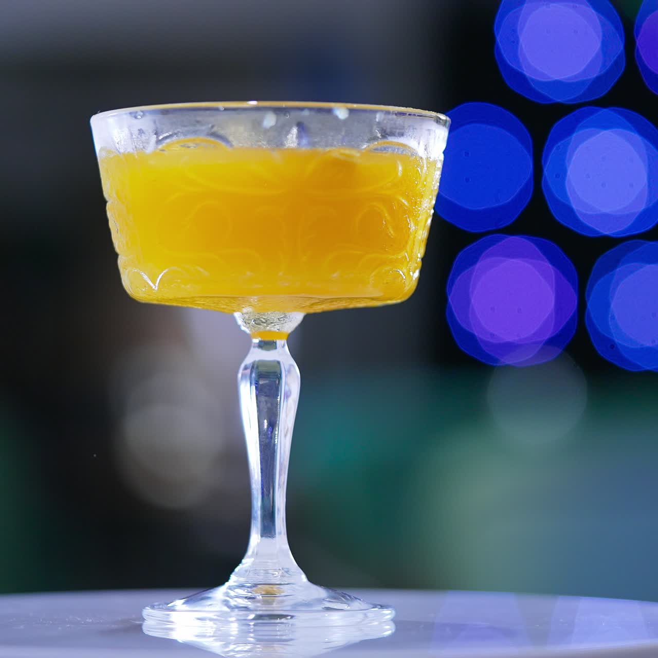 White rotating table with a glass of cocktail. Wide wineglass filled with orange beverage. Close up. Blue lights at backdrop in blur