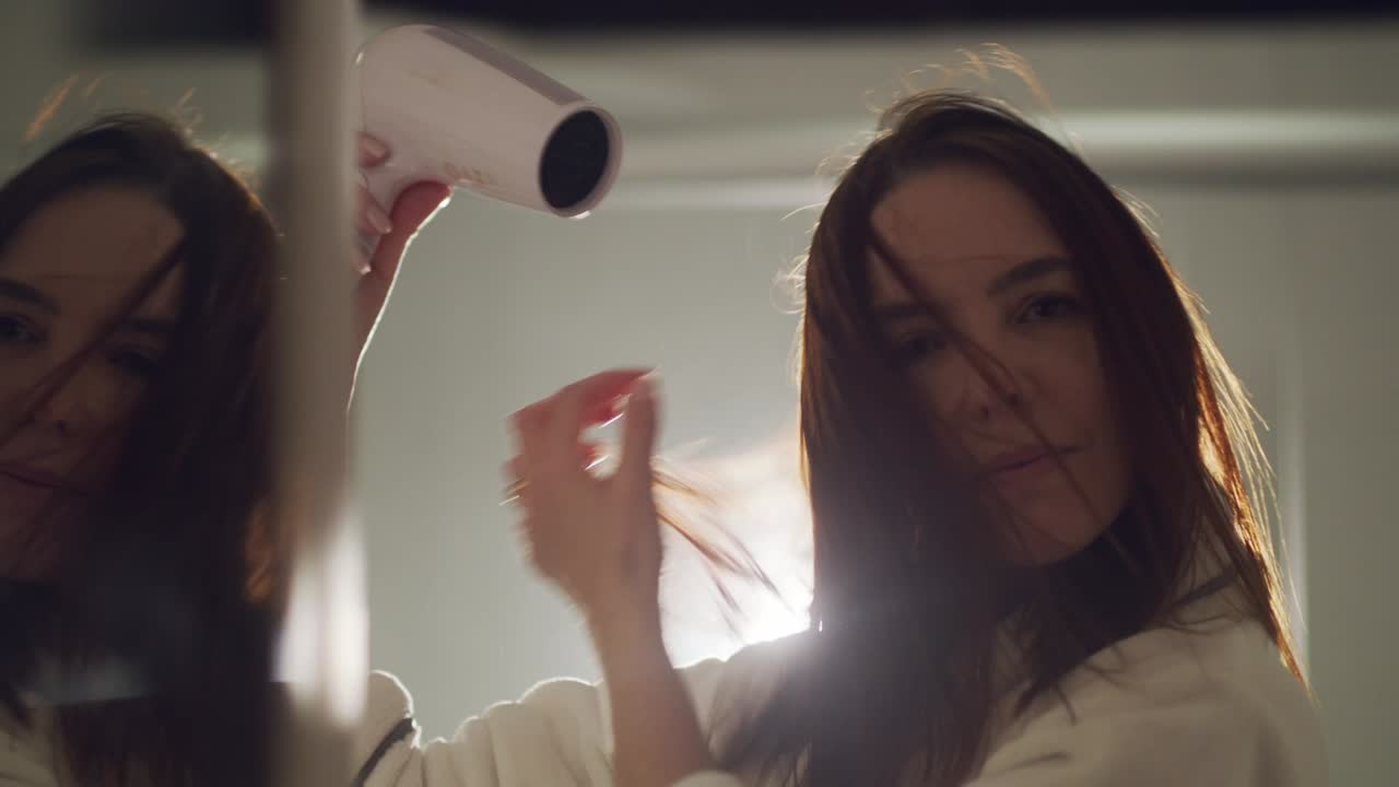 Brunette woman blow drying hair in bathroom, looking directly in camera
