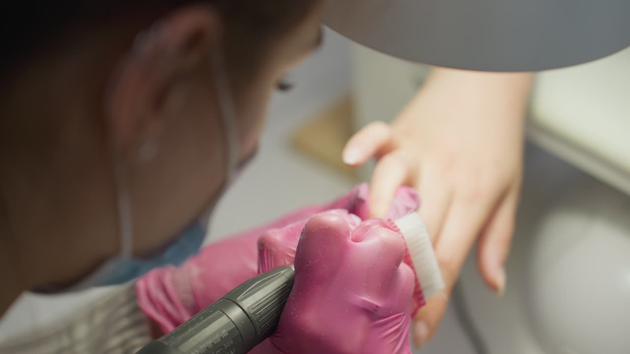 Top angle close up of nail technician wearing pink gloves and protective mask using brush and electric file to clean and shape client's nail during manicure session in modern salon environment
