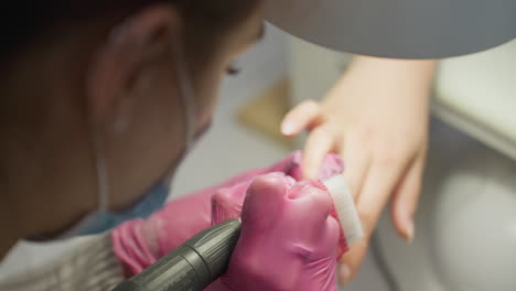 top angle close up of nail technician wearing pink gloves and protective mask using brush and electric file to clean and shape client's nail during manicure session in modern salon environment