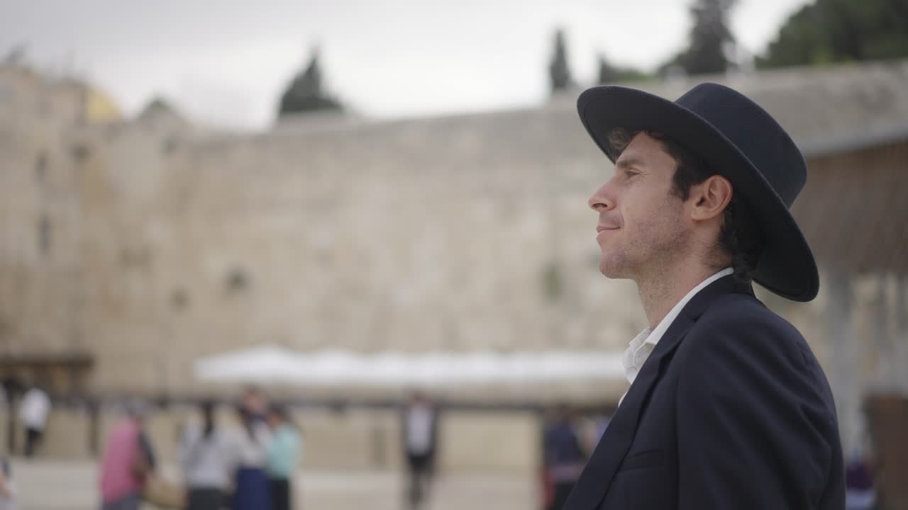 Jewish religious man at the Western Wall, Jerusalem, Israel. He walks in black suit and black hat in front of the wall in selective focus and slow motion.