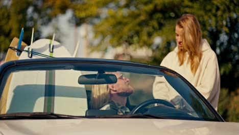 Ein-Blondes-Mädchen-In-Einem-Weißen-Sweatshirt-Kommuniziert-Mit-Ihrem-Blonden-Freund-Mit-Bart-Und-Brille,-Der-In-Einem-Weißen-Cabrio-Sitzt-Und-Auf-Dem-Zweiten-Sitz-Surfbretter-In-Der-Stadt-Vor-Kleinen-Bäumen-Mit-Grünem-Laub-Hat