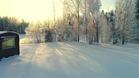 aerial drone panning shot over rectangular cottage and barrel sauna cabin on a snow covered white landscape during sunny morning