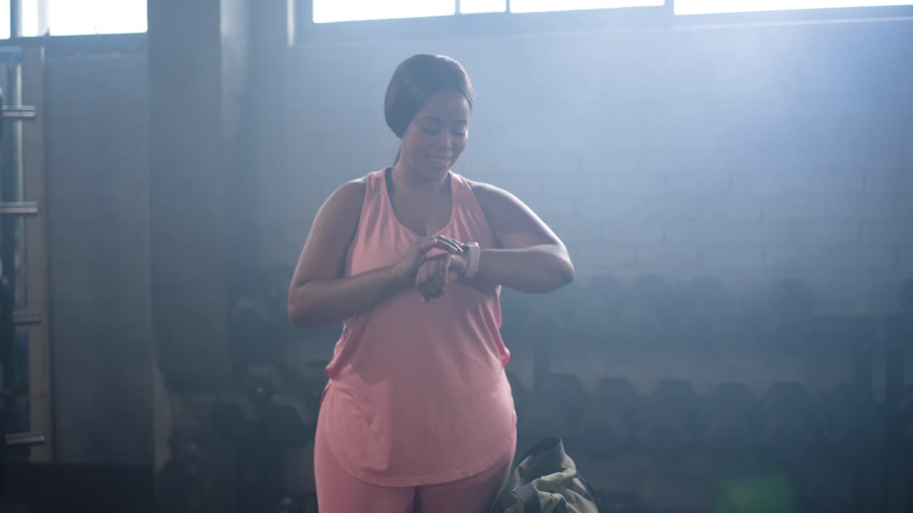 Donna afroamericana che entra in palestra con una bottiglia e una borsa in mano e controlla lo smartwatch mentre si prepara per l'allenamento