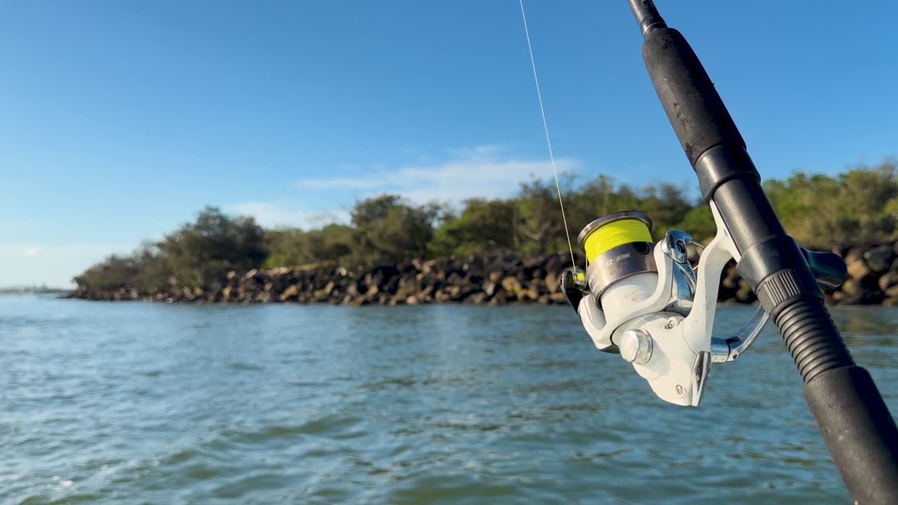 Primo piano di canna da pesca e mulinello con filo giallo, barca alla deriva su un estuario soleggiato