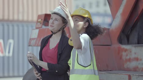 multi ethnic woman worker with hard helmet standing, working in container loading yard, checking and prepare container freight plan together. diverse employees and colleague in transport business.