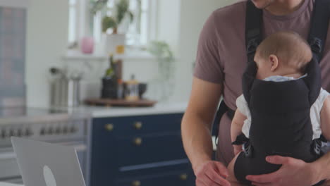 Primer-Plano-De-Un-Padre-Transgénero-Que-Trabaja-Desde-Casa-Con-Una-Computadora-Portátil-Cuidando-A-Su-Hijo-Dormido-En-Un-Cabestrillo