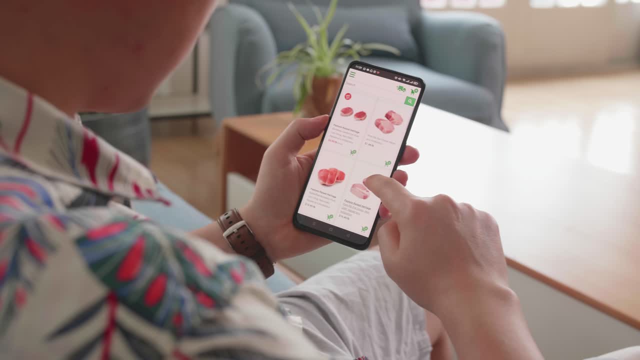 Man Sitting On Couch Using Smartphone Buys In Internet Shop. Male Selects Beef In Grocery Online Store