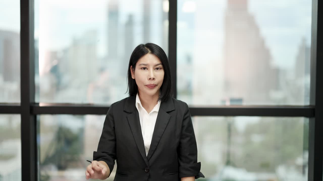 Asian businesswoman in suit smiles, makes OK gesture, natural daylight, modern office background
