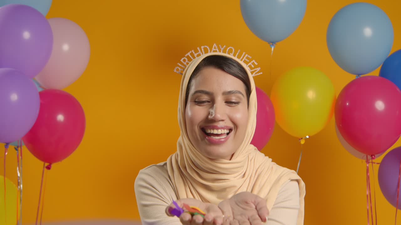Studio Portrait Of Woman In Hijab Wearing Birthday Queen Headband Celebrating Blowing Paper Party Confetti To Camera