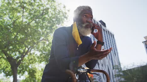 Afroamerikanischer-älterer-Mann-Mit-Gesichtsmaske,-Der-Auf-Dem-Smartphone-Spricht,-Während-Er-Sich-Auf-Sein-Fahrrad-Stützt