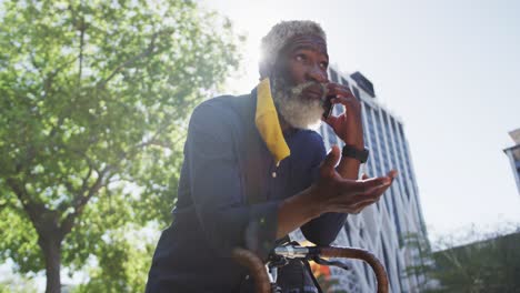 hombre mayor afroamericano con máscara facial hablando en un teléfono inteligente mientras se apoya en su bicicleta en