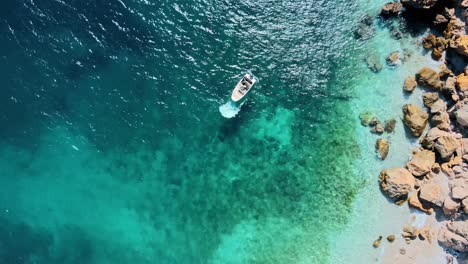 Skiff-or-boat-reverses-in-clear-turquoise-ocean-water-by-rocky-coast,-aerial-zenithal-view