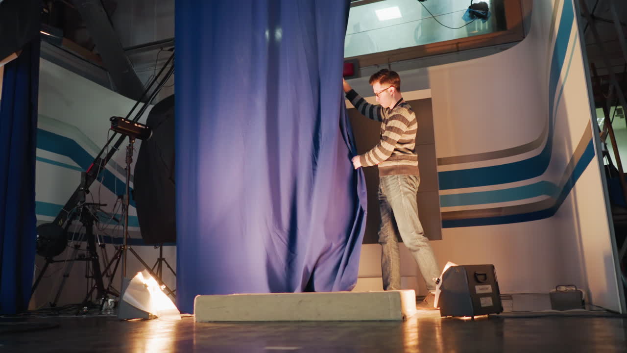 Young male stagehand pulls blue curtain during television studio setup, floor spotlight casts shadow on striped backdrop while crane camera gear and cables visible backstage broadcast production environment
