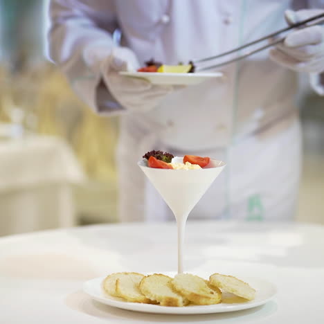 a waiter serves cottage cheese in a high ceramic bowl laying out cherry tomatoes, greens and lemon slices on the background of a white plate with five round slices of bread on the table in the restaurant.